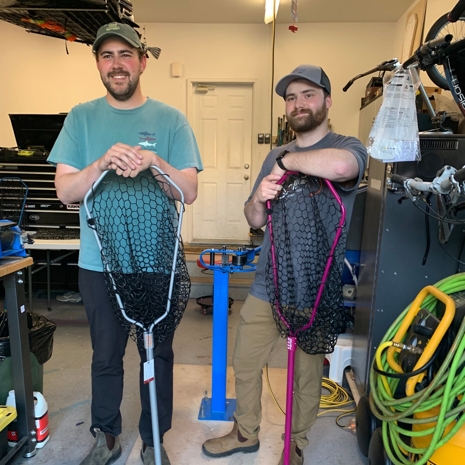 Two people standing in a garage or workshop with tools and equipment around holding landing nets.